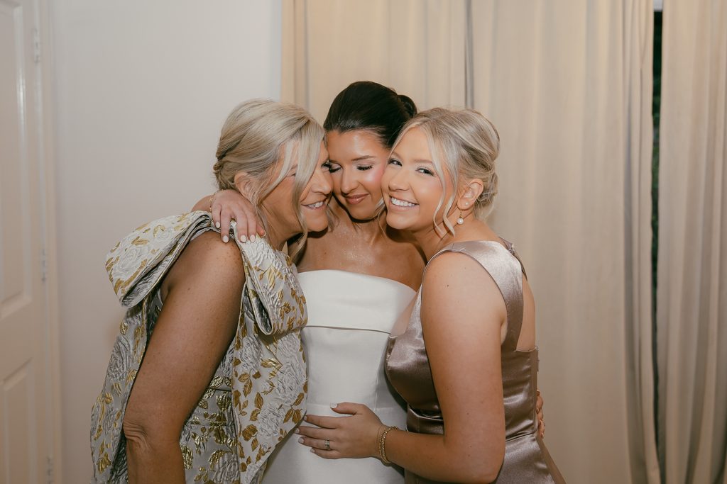 bride with her mom and sister pose closely together in their dresses, sharing a candid moment with the bride and bridesmaids.