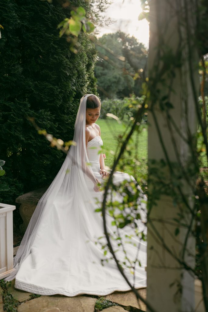 A bride in a white gown sits gracefully outdoors in a lush, green venue, highlighting a bride sitting down in a garden.